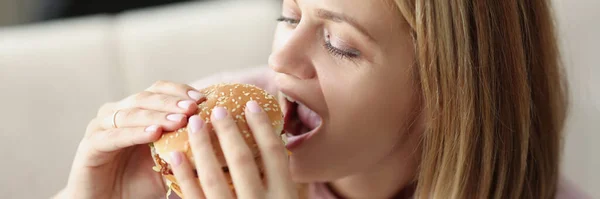 Jovem comendo delicioso hambúrguer apetitoso em casa — Fotografia de Stock