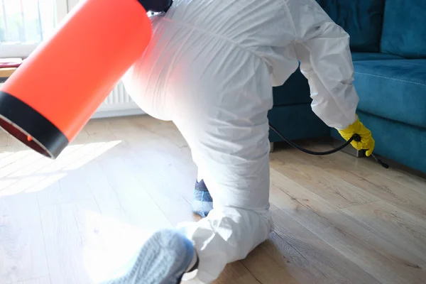 Travailleur en combinaison de protection traitant avec une solution désinfectante sur le sol sous le canapé dans l'appartement — Photo