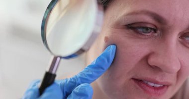 Eldivenli doktor, kadının yanağındaki beni büyüteçle inceliyor. Cildiyeci hastanedeki randevusunda hastanın cildindeki doğum lekesini kontrol etti.