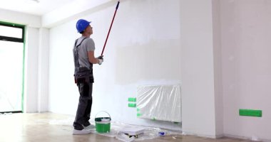 İnşaat işçisi, duvarın kaba yüzeyinde yavaşça ve düzenli bir şekilde yuvarlanıyor. Miğferli eleman duvara parlak beyaz boya sürüyor.