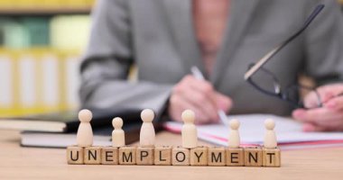 Sessiz bir çalışma alanında, bir kişi işsizlikle ilgili bir belgeyi incelerken, ön planda tahtadan fayanslar UNEMPLOYMENT sözcüğünü heceler. Atmosfer yansımayı ve endişeyi yansıtır..