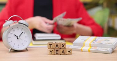 Vergi hazırlamakla ilgili bilinen eşyalar düzenli bir çalışma alanını sararken elleri para sayarken görülüyor. Bir çalar saat, tamamlanan mali göreve aciliyet katar.