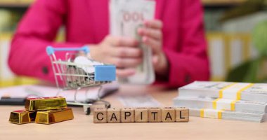 Elleri para saymakla ve altın külçelerini düzenlemekle meşgulken harf blokları sermaye kelimesini heceliyor. Atmosfer canlı, akıllı yatırımların ve zenginlik yönetiminin önemini gösteriyor..