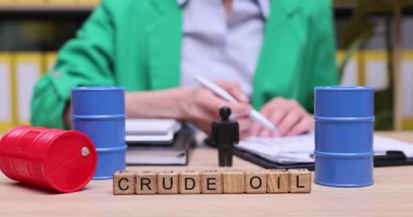 Yeşil ceketli bir kişi ham petrol ticareti hakkında veri ve trendler üzerinde çalışıyor. Kırmızı ve mavi variller piyasa dinamiklerini gözler önüne sererken, ahşap bloklar ham petrolü hecelerken, anahtar kavramları yansıtıyor..