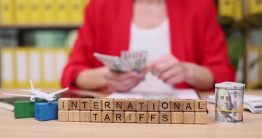 Renkli dosyalarla dolu canlı bir alanda, bir kişi uluslararası tarifelerle etiketlenmiş ahşap blokları düzenlerken nakit parayı inceliyor. Minyatür uçak ve konteyner modelleri yakınlarda..