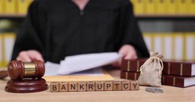 Siyah bornozlu bir kişi, iflas sürecini açıklayan evrak işleri ve yasal eşyalarla etkileşime giriyor. Gavel ve tahta bloklar belirgin bir şekilde, ciddi bir yasal ortamı vurguluyor..