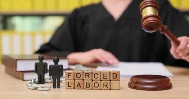 Bir hakim zorla çalıştırılan işçilik sorununa, zorunlu işçilik kelimelerini içeren tahta bloklarla değiniyor. Mahkeme salonu ciddi bir hava sağladığı için önemli yasal belgeler ele alınıyor..