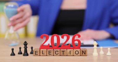 Satranç taşları ve canlı bloklar 2026 seçim temasını gözler önüne seriyor. Bir figür düşünceli bir şekilde satranç zamanlayıcısı kullanarak bir beklenti ve planlama atmosferi yaratıyor..