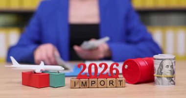 Bir kişi, 2026 yılında ithalat sektörünü büyütmek için yeni stratejileri simgeleyen renkli bloklar ve model bir uçakla çevriliyken para sayıyor. Yaratıcı görseller girişimci enerjiyi iletir.