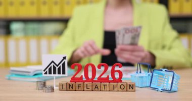 Parlak bir ofiste, kişi 2026 yılındaki enflasyon eğilimlerini analiz ederken mali belgeleri ve parayı inceler. Renkli tablolar ve simgeler masadaki ekonomik görüşleri gösteriyor.