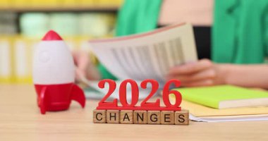 Kitaplarla dolu parlak bir alanda, eller renkli belgelerin sayfalarını çeviriyor. Yenilikçi ruhun ve değişikliklerin 2026 'da gerçekleşeceğinin sinyallerini veren oyuncu bir roket oyuncağı yakınlarda duruyor..