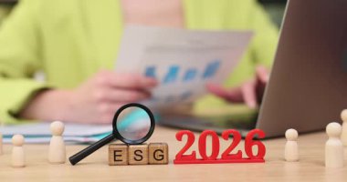 Parlak bir ofiste, eller sürdürülebilirliğe odaklı çizelge ve stratejileri inceler. Kırmızı harfler 2026 yazarken, büyüteç ESG ilkelerinin önemini vurguluyor..