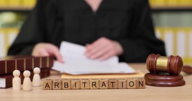 Bir yargıç masasında otururken hakemlik belgelerini dikkatle inceler. Yanlarında, tahta bloklar ARBITRation 'ı heceliyor ve küçük rakamlar katılımcıları temsil ediyor.
