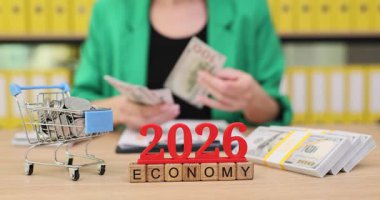 Bir kişi bir masada oturur, nakit para yığınları ve küçük bir alışveriş arabasıyla birlikte parayı sayar. Projede 2026 yılına kadar ekonomik iyileşme potansiyeli üzerinde duruluyor..