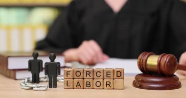 Bir mahkeme salonunda, bir yargıç zorla çalıştırma sorununa değinir. Tahta bloklar zorunlu işçiliği ifade ederken, tokmak bu önemli sosyal konunun aciliyetini vurgular..