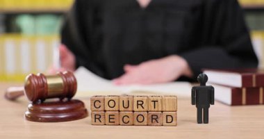 Bir yargıç masada oturup önemli mahkeme kayıtlarını inceliyor. Tokmak ve diğer yasal eşyalar yakınlarda dinlenirken onun dikkati evrak işlerinde. Ciddi bir atmosfer yaratıyor..