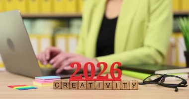 2026 için yaratıcı fikirler planlamaya odaklanan parlak yeşil ceketli bir kişi, renkli kırtasiye ve gözlüklerle çevrili bir dizüstü bilgisayarda yazı yazar..