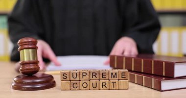 Bir yargıç, kitapların yanındaki belgelere odaklanarak, resmi bir mahkeme salonunda önemli bir davaya hazırlanır. Bir tokmak masada duruyor, otoriteyi ve adaleti simgeliyor..