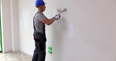 Miğferli ressam apartman onarımı sırasında duvara beyaz boya sürüyor. İşçi ofis renk temasını geniş muhafaza yavaş çekimde yeniliyor