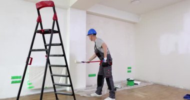 Üniformalı müteahhit dairede merdivenin yanında duran duvarı beyaza boyuyor. İşçi, evin içini tekerlekli fırça kullanarak dekore ediyor.