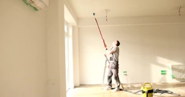 Üniformalı adam tavanı beyaz renge boyuyor ve evde paten fırçası var. İnşaatçı dairede ışık pigmenti ile iç tasarım tarzını korur