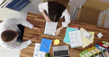 Erkek yönetici, yönetim kurulundaki kadın ortağa sözleşmeli pano veriyor. İş arkadaşları toplantıda resmi evraklarla ilgilenirler.