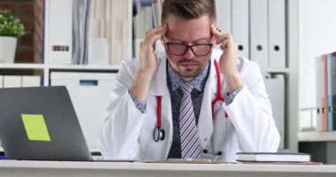 Gözlüklü doktor masa başında oturup dizüstü bilgisayarın yanındaki şakaklara bastırıyor. Doktor, teşhis planını göz önünde bulundurarak evrak işlerinden dolayı stres altında.