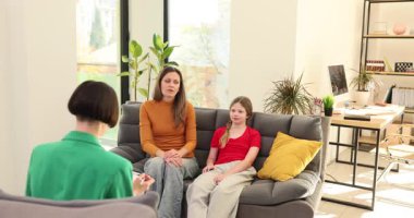 Asyalı terapist, rahat oturma odasında beyaz anne çocuğunu dinliyor. Yeşil ceketli klinisyen, annesi ve kızıyla gri giyinmiş.