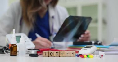 Tahta bloklar ICD-11 'i işaret ediyor burun spreyi monitörünün yanında ve masada doktor aleyhinde haplar var. Kadın hekim, ICD kurallarıyla x-ray güncelleme tanısını gözden geçirdi