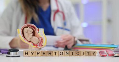Wooden blocks spell word Hypertonicity near uterus model fetus and stethoscope on table. Woman obstetrician writes notes evaluating uterine tone