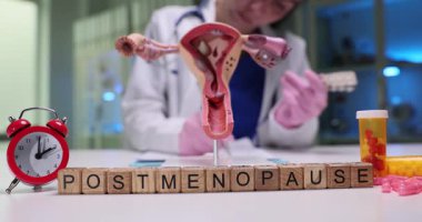 Rahmin yakınındaki tahta küplerde menopoz sonrası cümleler ve klinikteki alarm saati ve haplar. Jinekolog tedavi için hasta kayıtlarını dolduruyor.