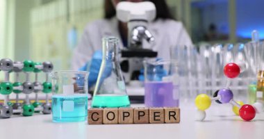 Lab 'da renkli sıvıların ve moleküler yapı modellerinin yanındaki tahta bloklardaki bakır kelime. Kadın kimyager, masada mikroskop kullanarak metali araştırıyor.