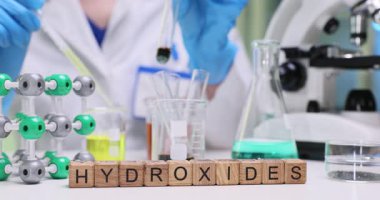 Genç kadın kimyager yavaş çekimde sıvıya reaktif madde katıyor. Laboratuvar masasında minyatür ahşap küpler, hidroksit terimi.