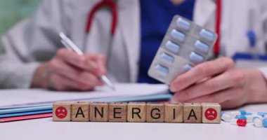 Masa yüzeyine yerleştirilmiş minyatür tahta küpler Anergia kelimesini oluşturuyor. Doktor reçete yazıyor ve elinde bir paket hap tutuyor.