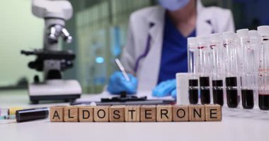 Tahta bloklar kan test tüplerinin yanında Aldosteron kelimesini heceler. Laboratuvar doktoru hasta örneklerini mikroskop ile inceleyen detaylı hormon raporu yazıyor.