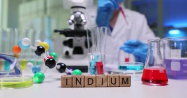 Tahta bloklar laboratuardaki parlak kimya masasında Indium kelimesini heceler. Beyaz önlüklü bilim adamı metalin özelliklerini inceleyen renkli çözümleri karıştırıyor