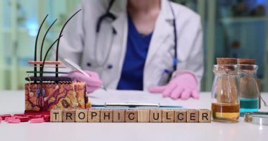 Tahta bloklar tıbbi masadaki deri modelinin yanında Trofik Ülser olarak yazılıyor. Beyaz önlüklü doktor kadın masadaki yara tabakalarında notlar yazıyor.