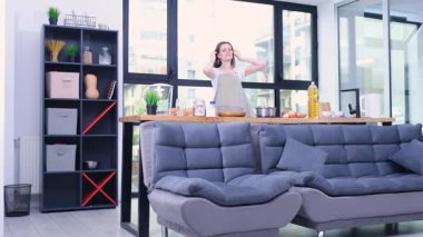 Gülümseyen kadın yemek yapmadan önce parlak bir ev mutfağında önlük giyer. Kadınlar aile yemeği yapmaya hazırlanırken masadaki malzemeler bekliyor.