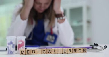 Minyatür tahta küpler tıbbi hata ifadesini oluşturur. Sıkıntılı kadın doktor masa başında oturur ve elleri şakaklara tutunur.