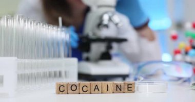 Kadın kimyager, mikroskop altında kokain tozunu dikkatlice inceliyor. Minyatür ahşap küpleri Kokain sözcüğünün masa biçiminde düzgünce düzenlenmiştir.