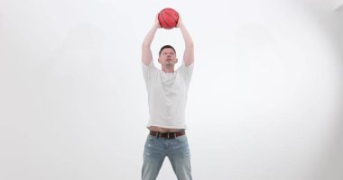 Adam basketbol topuyla beyaz arka planda ağır çekimde egzersiz yapıyor. Güçlü sporcu turuncu küreyi kaldırıyor ve ışık duvarının yakınına fırlatmaya hazırlanıyor.