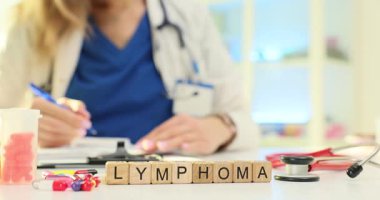 Tahta bloklar, masadaki dağınık hapların yanında Lenfoma kelimesini heceliyor. Onkoloji doktoru randevuda lenfoma aşaması ve terapiyi tartışan vaka notları yazar.