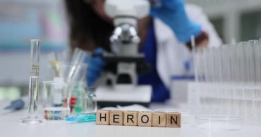 Tahta bloklar şırınganın yanındaki laboratuvar tezgahında eroin kelimesini heceler. Adli tıp uzmanı kadın, işyerindeki eroin örneklerini incelerken mikroskop kullanıyor.