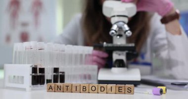 Tahta bloklar kan örnekleri ve tüplerin yanında antikor kelimelerini heceler. Laboratuvar kadını çalışmaları mikroskop altında antikor seviyesini ölçer.