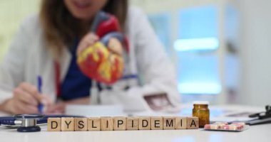 Tahta bloklar anatomik kalp modelinden Dyslipidemia kelimesini heceler. Kardiyoloji doktoru randevuda dispidemi tedavi planını açıklayan notlar yazar.