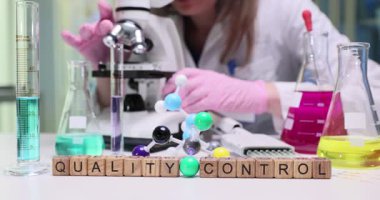 Laboratuvar teknisyeni numuneyi mikroskop altında dikkatlice inceliyor. Tablo biçimindeki tahta bloklar cam eşyalar ve ekipmanlar arasında Kalite Kontrolü