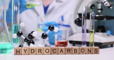 Kadın laboratuvar çalışanı yavaş yavaş canlı sarı çözeltiyi analiz aracına aktarıyor. Minyatür tahta küpler masada hidrokarbon terimi oluşturur. Yavaş çekim