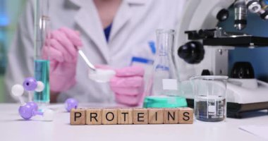 Laboratuvar çalışanı şeffaf kabın içinde ince beyaz toz karıştırıyor. Masadaki minyatür tahta küpler açıkça Protein kelimelerini oluşturuyor. Yavaş çekim