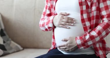 Kareli gömlekli hamile bir kadın kanepeye oturup iki eliyle karnını okşuyor. Anı, annelik ve aile planlamasını simgeler.