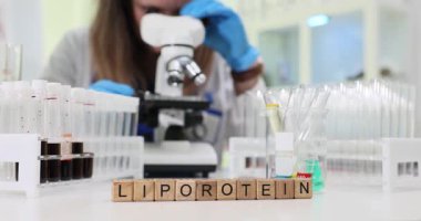 Tahta bloklar kelime Lipoprotein mikroskop yanında ve test tüpleri masada. Kadın doktor kan örneğini incelerken göz merceğine bakar.